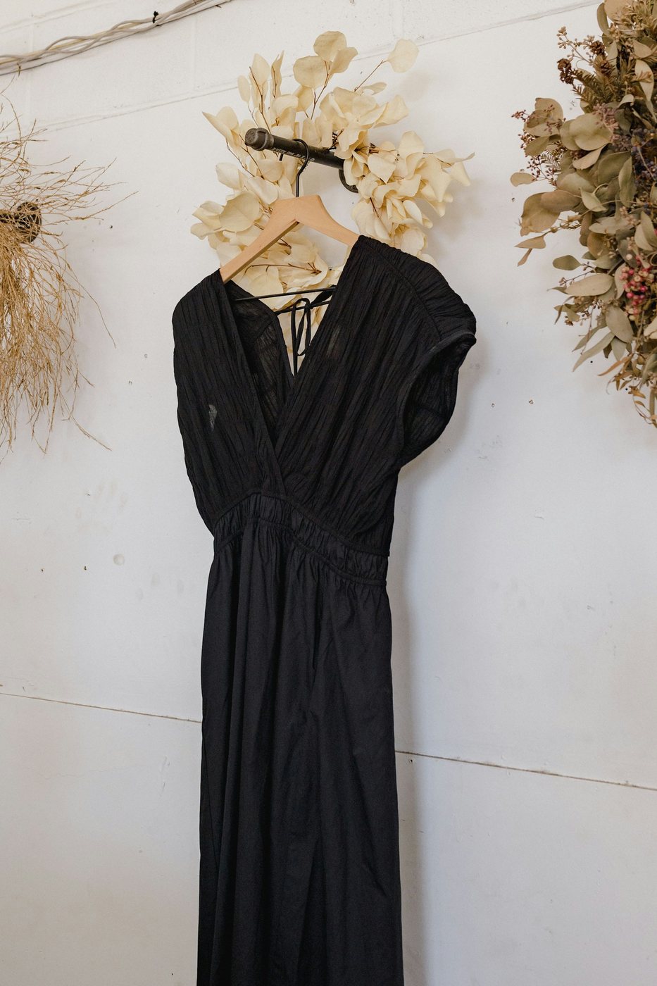 Black crossover dress on a wooden hanger against a white wall hung with dried wreaths.