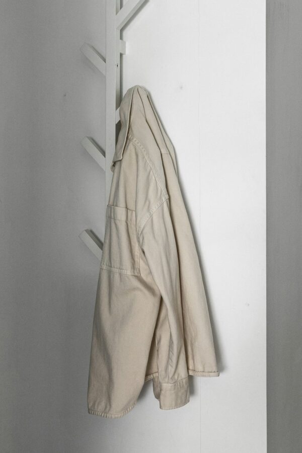 Ecru canvas overshirt hanging from a white wall hook.