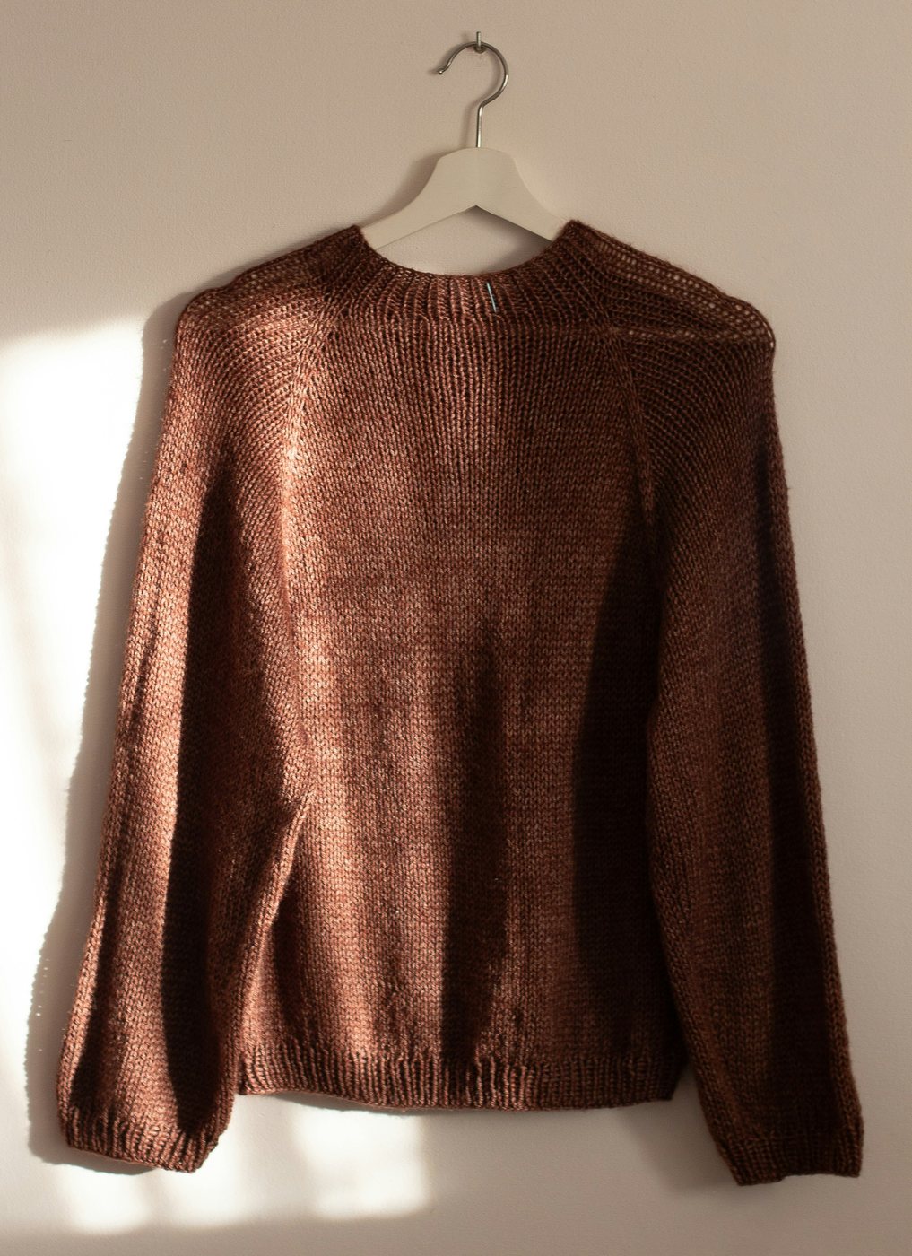 Chestnut brown knit sweater on a white hanger against a sunlit wall.