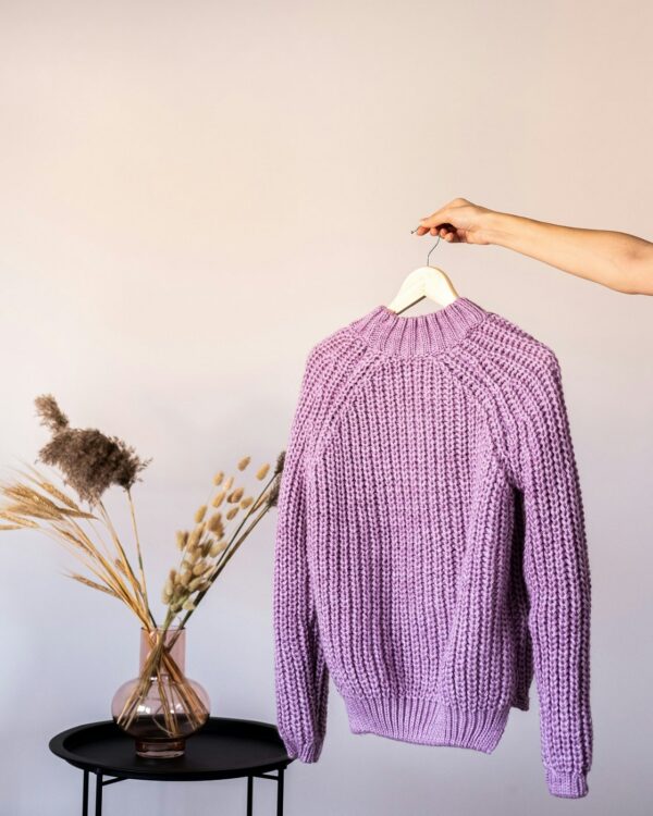 Lilac chunky rib sweater held up on a hanger next to a vase of dried grasses.
