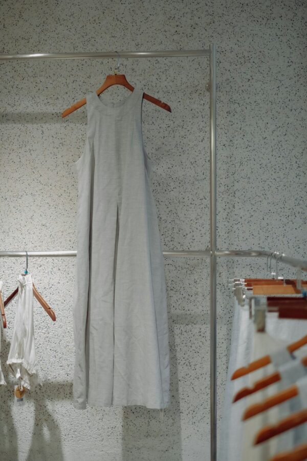 Grey sleeveless ankle-length linen dress on a wooden hanger against a speckled wall.