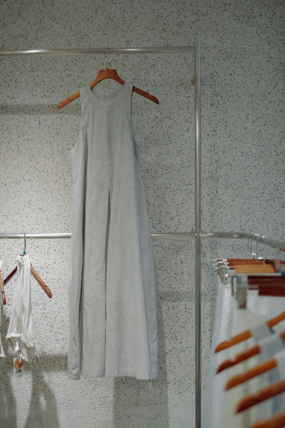Grey sleeveless ankle-length linen dress on a wooden hanger against a speckled wall.