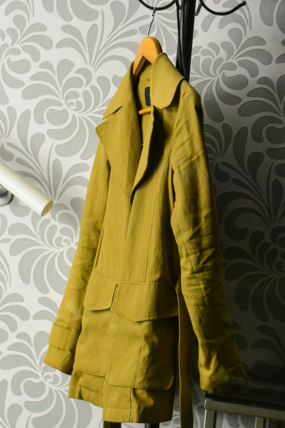 Mustard field jacket on a wooden hanger on a coat stand in front of patterned wallpaper.