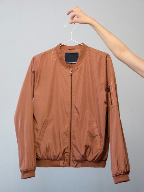 Rust bomber jacket held up on a white hanger against a grey wall.