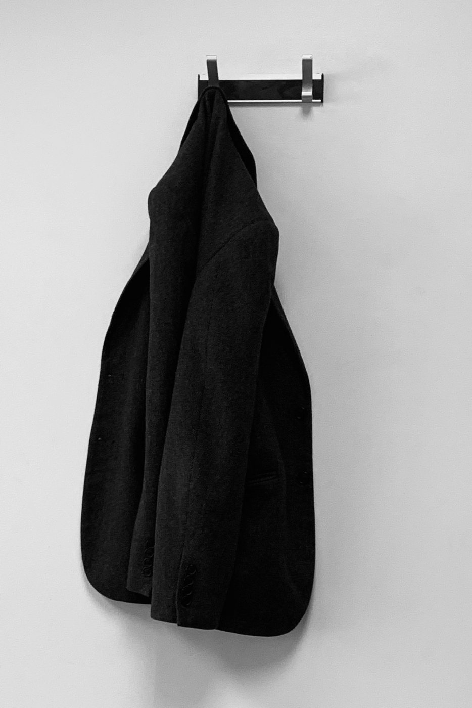 Black wool blazer hanging from a hook on a white wall.